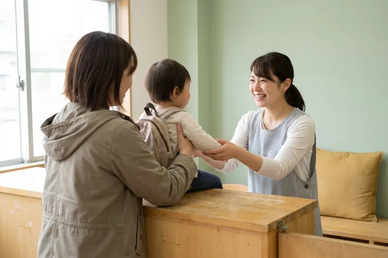 A parent handing a toddler with a small backpack to a caregiver at a bright daycare front counter